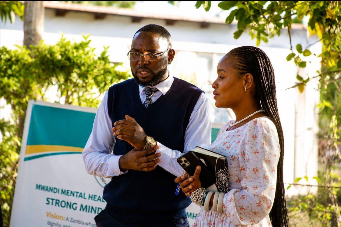 Patrick Mwewa in conversation at the Mwandi outreach, a banner reading Strong Minds, Strong Zambia behind him